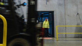 Mies astuu ulos tehdashallin ovesta yllään turvavaatetus. Trukki ajaa ohi. Mies astuu ulos tehdashallin ovesta yllään turvavaatetus. Trukki ajaa ohi.