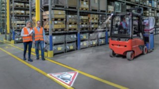 Two people walk in the warehouse and cross paths with a forklift truck
